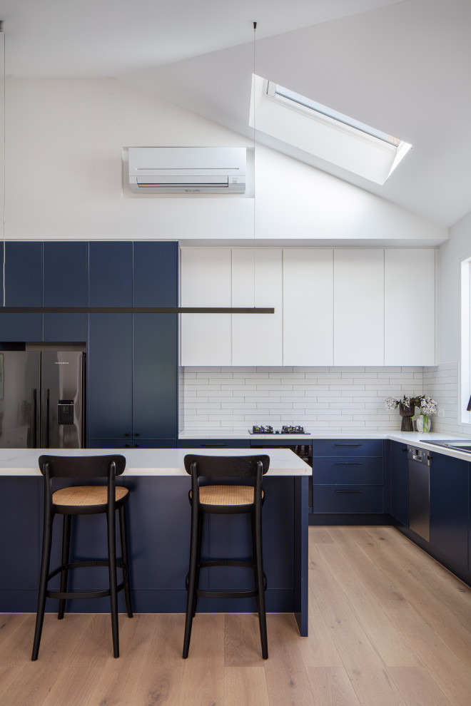 Design ideas for a contemporary l-shaped kitchen with a drop-in sink, blue cabinets, white splashback, stainless steel appliances, light hardwood floors, with island, beige floor, white benchtop and vaulted.