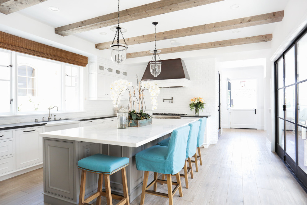 Large beach style l-shaped kitchen in Orange County with a farmhouse sink, shaker cabinets, white cabinets, marble benchtops, white splashback, stainless steel appliances, light hardwood floors and with island.