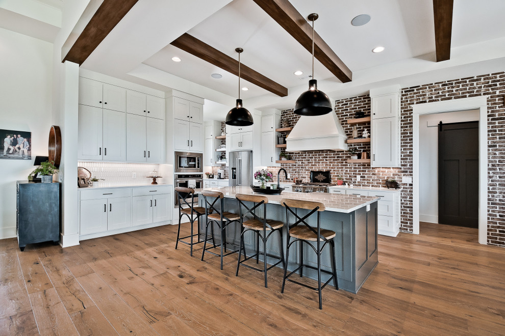 Photo of a large farmhouse u-shaped kitchen/diner in Other with a belfast sink, raised-panel cabinets, white cabinets, engineered stone countertops, red splashback, brick splashback, stainless steel appliances, light hardwood flooring, an island, white worktops and exposed beams.