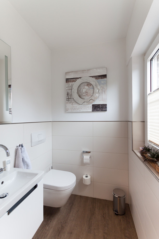 Inspiration for a small beach style powder room in Bremen with flat-panel cabinets, white cabinets, a wall-mount toilet, beige tile, ceramic tile, white walls, vinyl floors, an integrated sink and brown floor.