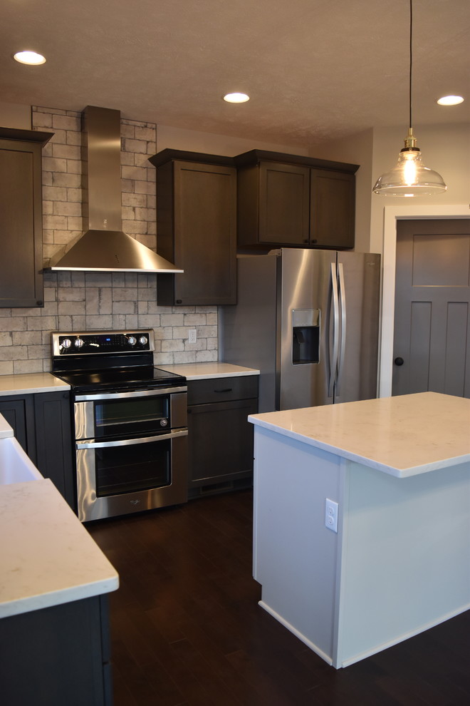 Photo of a mid-sized country l-shaped kitchen in Other with a farmhouse sink, recessed-panel cabinets, grey cabinets, grey splashback, brick splashback, stainless steel appliances, dark hardwood floors and with island.