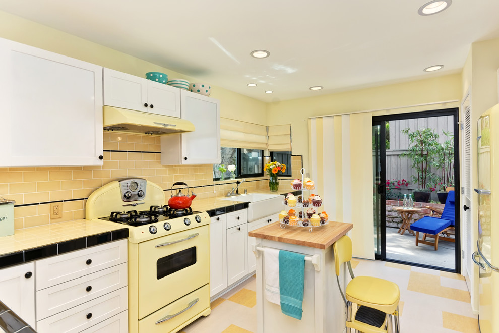 This is an example of a mid-sized traditional l-shaped eat-in kitchen in San Diego with a farmhouse sink, shaker cabinets, white cabinets, tile benchtops, yellow splashback, subway tile splashback, coloured appliances, linoleum floors and a peninsula.