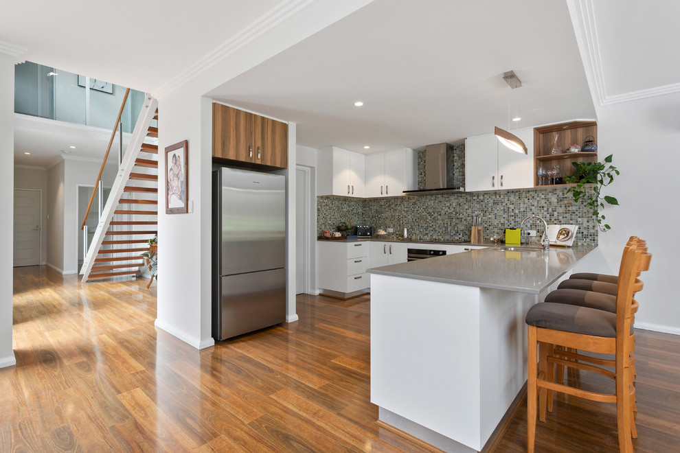 Inspiration for a transitional u-shaped kitchen in Perth with an undermount sink, flat-panel cabinets, white cabinets, grey splashback, mosaic tile splashback, stainless steel appliances, medium hardwood floors, brown floor and grey benchtop.