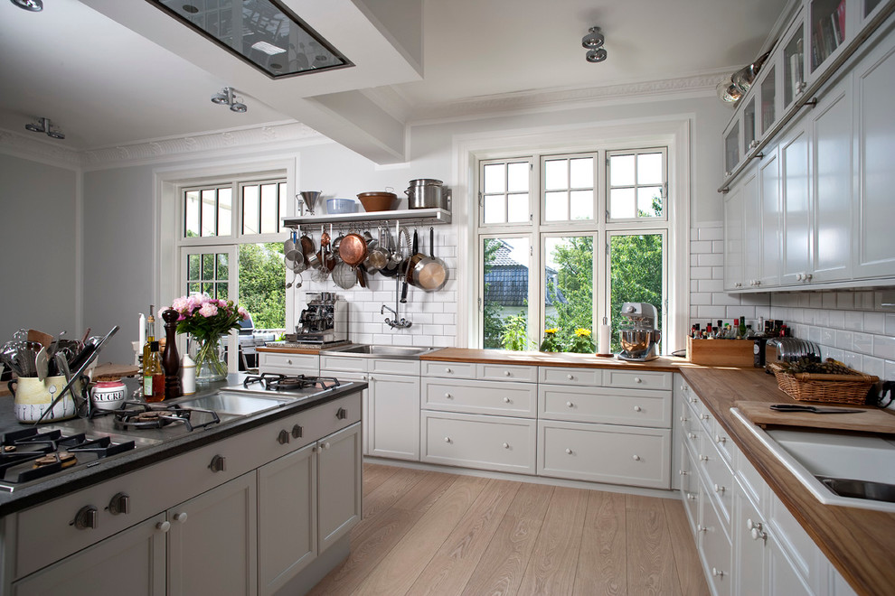 Photo of a large traditional u-shaped open plan kitchen in Aalborg with a drop-in sink, recessed-panel cabinets, white cabinets, wood benchtops, white splashback, subway tile splashback and light hardwood floors.