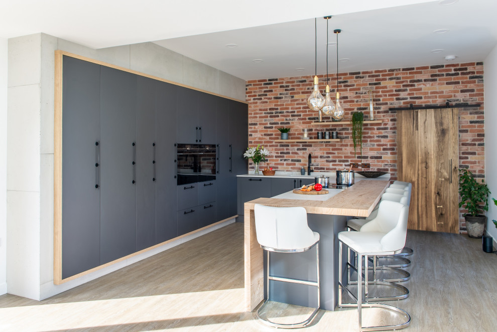 This is an example of a large modern u-shaped open plan kitchen in Cornwall with a drop-in sink, flat-panel cabinets, grey cabinets, multi-coloured splashback, brick splashback, black appliances, light hardwood floors, with island and white benchtop.