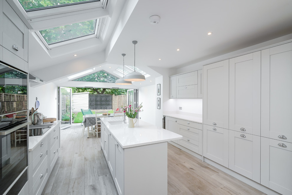 Inspiration for a mid-sized transitional galley eat-in kitchen in London with recessed-panel cabinets, solid surface benchtops, porcelain floors, with island, white benchtop, an undermount sink, white cabinets, white splashback and beige floor.
