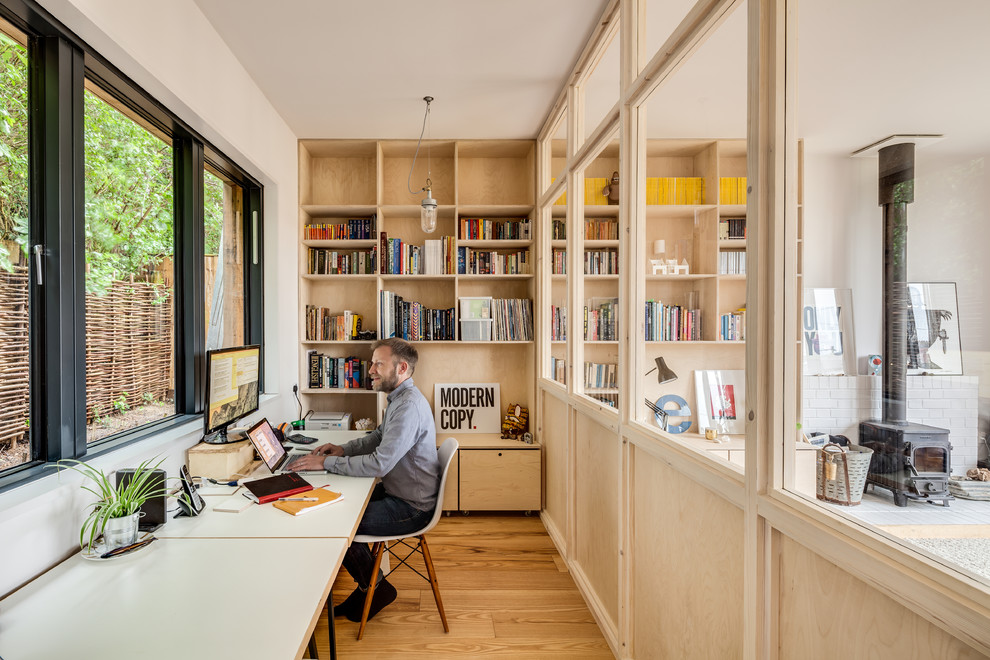 Mid-sized scandinavian home studio in Buckinghamshire with white walls, light hardwood floors, a wood stove, a brick fireplace surround, a built-in desk and brown floor.