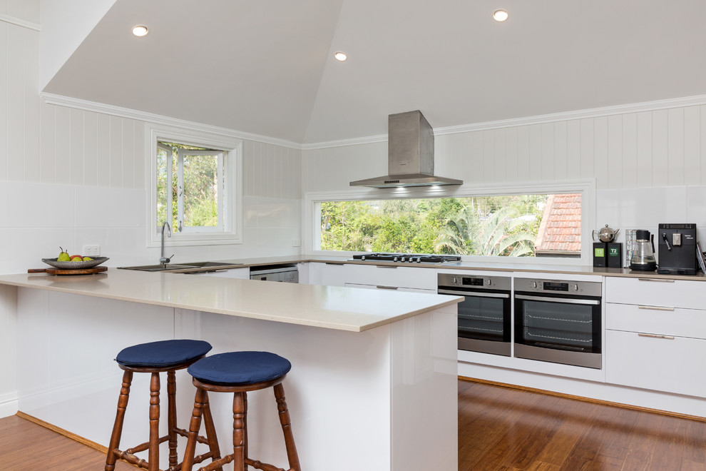 Inspiration for a mid-sized beach style u-shaped separate kitchen in Brisbane with a drop-in sink, flat-panel cabinets, quartz benchtops, white splashback, stainless steel appliances, bamboo floors and with island.