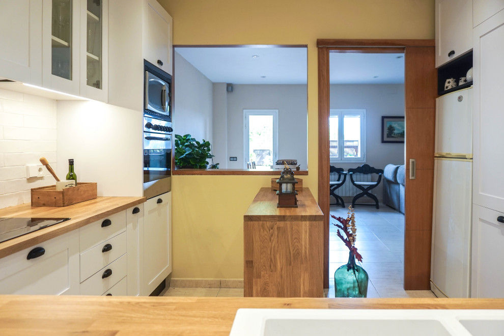 バルセロナにある低価格の中くらいなラスティックスタイルのおしゃれなキッチン (ダブルシンク、ガラス扉のキャビネット、白いキャビネット、木材カウンター、白いキッチンパネル、セラミックタイルのキッチンパネル、白い調理設備、セラミックタイルの床、ベージュの床、茶色いキッチンカウンター) の写真