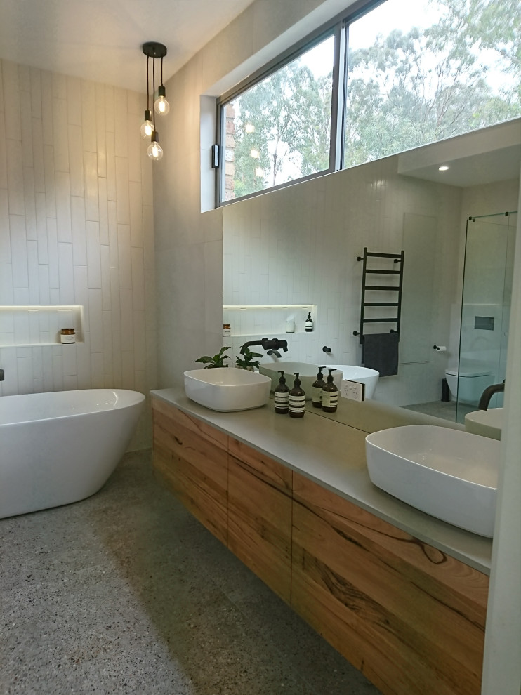 This is an example of a large modern bathroom in Melbourne with flat-panel cabinets, medium wood cabinets, a freestanding tub, a one-piece toilet, white tile, ceramic tile, white walls, terrazzo floors, a vessel sink, engineered quartz benchtops, grey floor, a hinged shower door, a niche, a double vanity, a floating vanity, vaulted and grey benchtops.