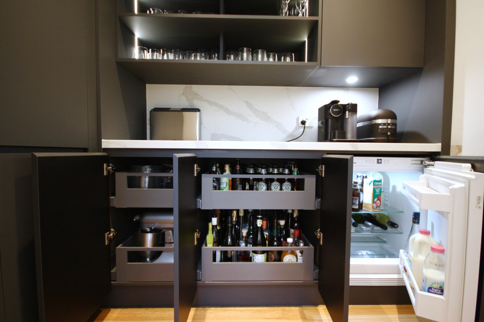 This is an example of a large modern single-wall open plan kitchen in Sydney with a submerged sink, flat-panel cabinets, grey cabinets, engineered stone countertops, white splashback, engineered quartz splashback, black appliances, medium hardwood flooring, an island, brown floors and white worktops.