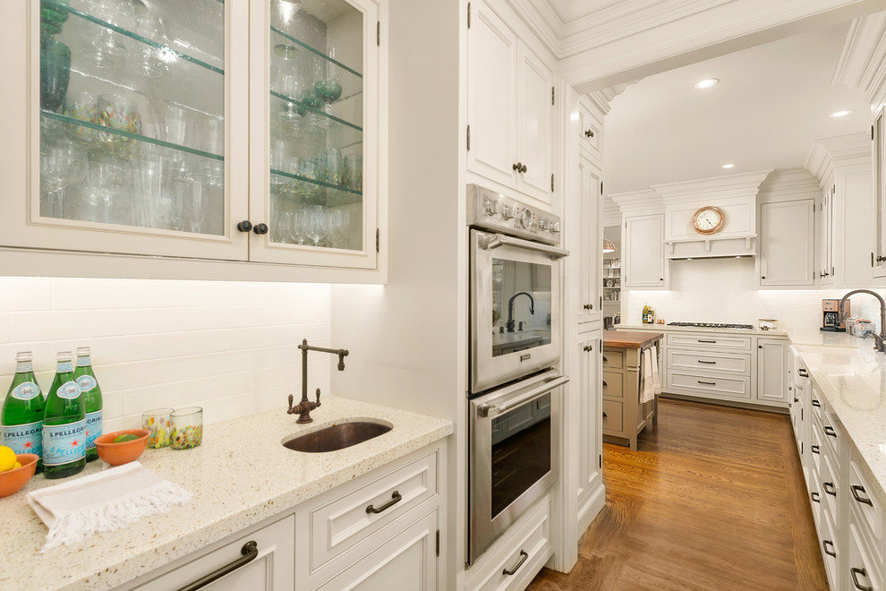 Inspiration for a mid-sized mediterranean l-shaped open plan kitchen in San Francisco with a farmhouse sink, recessed-panel cabinets, white cabinets, terrazzo benchtops, white splashback, subway tile splashback, panelled appliances, medium hardwood floors, with island, brown floor and beige benchtop.