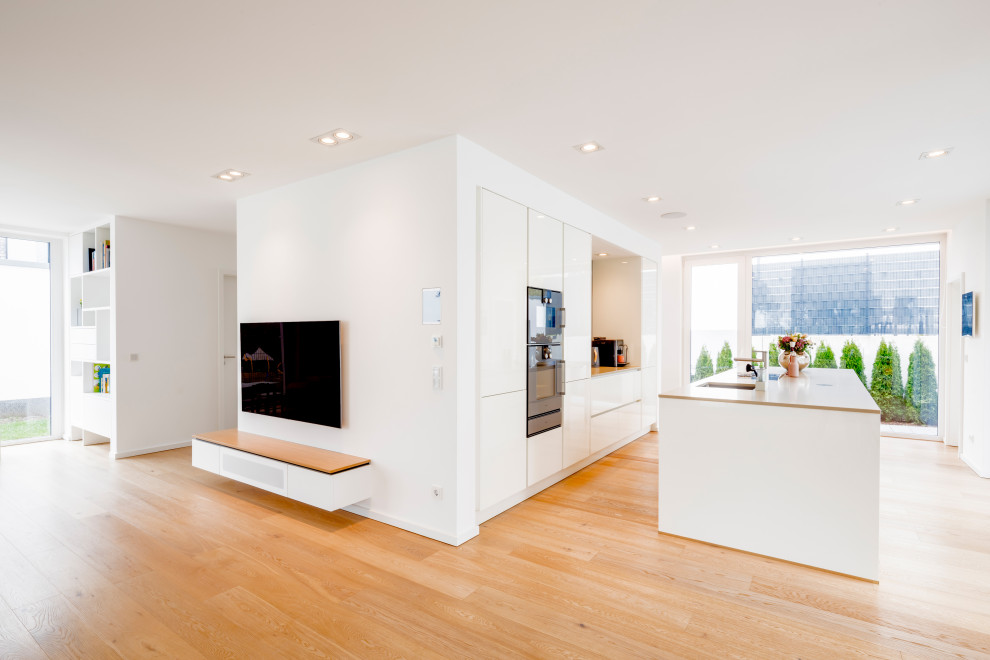 Inspiration for a large contemporary single-wall open plan kitchen in Dusseldorf with a built-in sink, flat-panel cabinets, white cabinets, quartz worktops, grey splashback, black appliances, medium hardwood flooring, an island, brown floors and beige worktops.