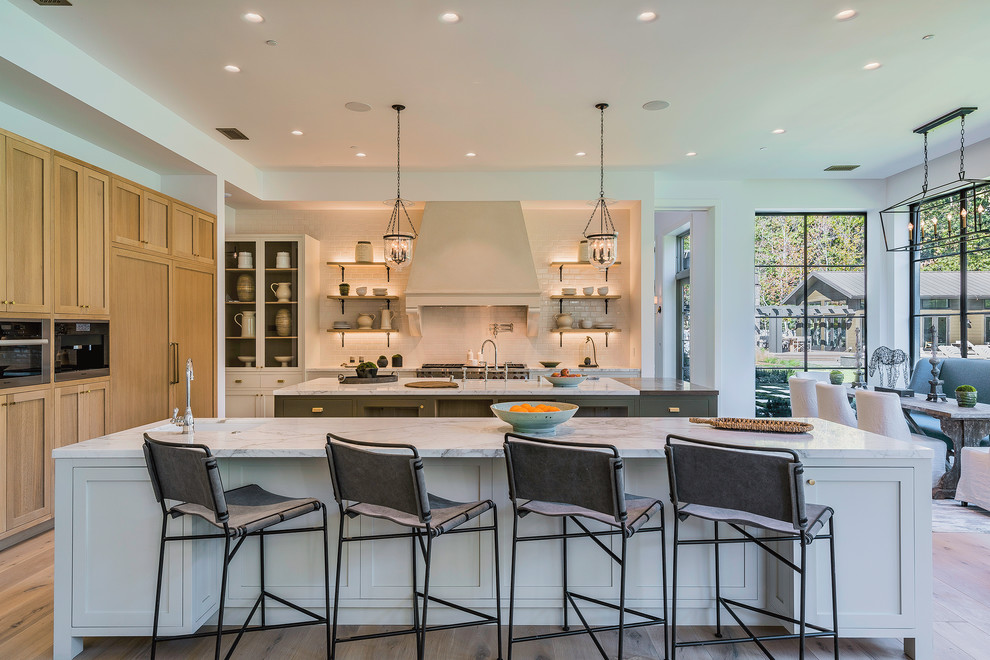 Large country l-shaped eat-in kitchen in Los Angeles with marble benchtops, white splashback, subway tile splashback, light hardwood floors, multiple islands, beige floor, a farmhouse sink, recessed-panel cabinets, light wood cabinets and stainless steel appliances.
