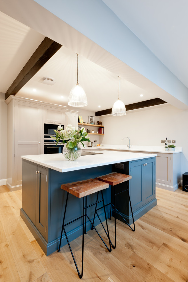 Mid-sized transitional l-shaped kitchen in Kent with shaker cabinets, white cabinets, white splashback, panelled appliances, light hardwood floors, with island, beige floor and white benchtop.