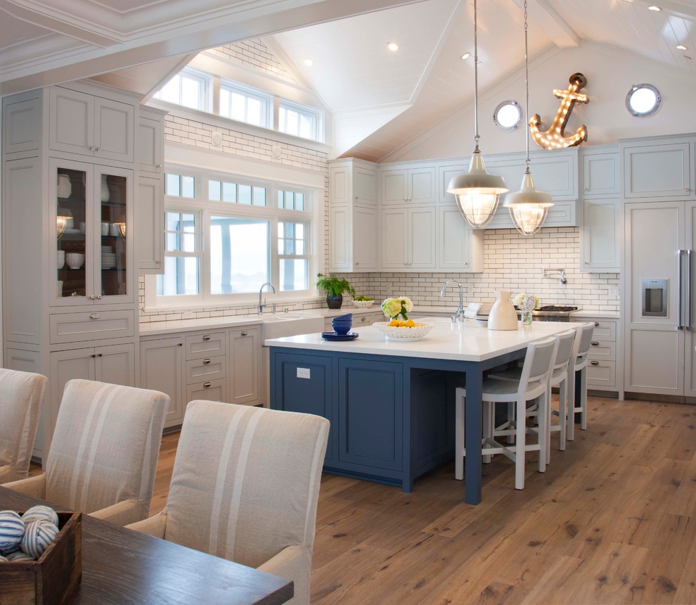 Photo of a beach style l-shaped eat-in kitchen in San Diego with a farmhouse sink, recessed-panel cabinets, white cabinets, white splashback, subway tile splashback, panelled appliances, medium hardwood floors, with island and brown floor.