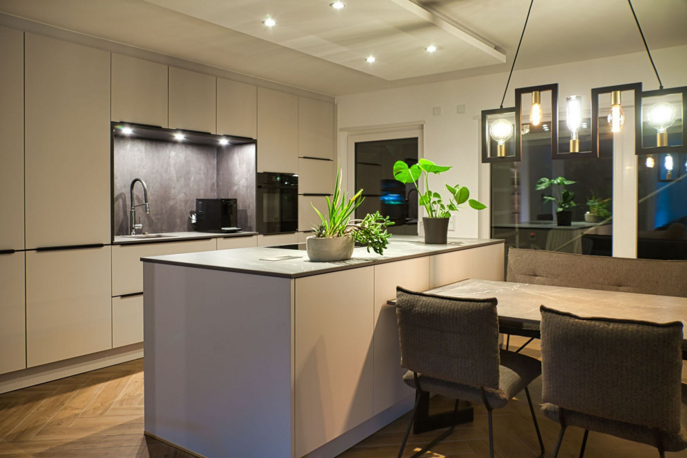 This is an example of a mid-sized contemporary galley open plan kitchen in Other with a drop-in sink, flat-panel cabinets, white cabinets, solid surface benchtops, grey splashback, black appliances, painted wood floors, with island, brown floor, grey benchtop and wallpaper.