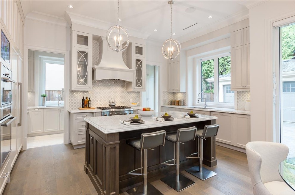 This is an example of a transitional u-shaped kitchen in Vancouver with an undermount sink, shaker cabinets, white cabinets, white splashback, stainless steel appliances, medium hardwood floors, with island and brown floor.