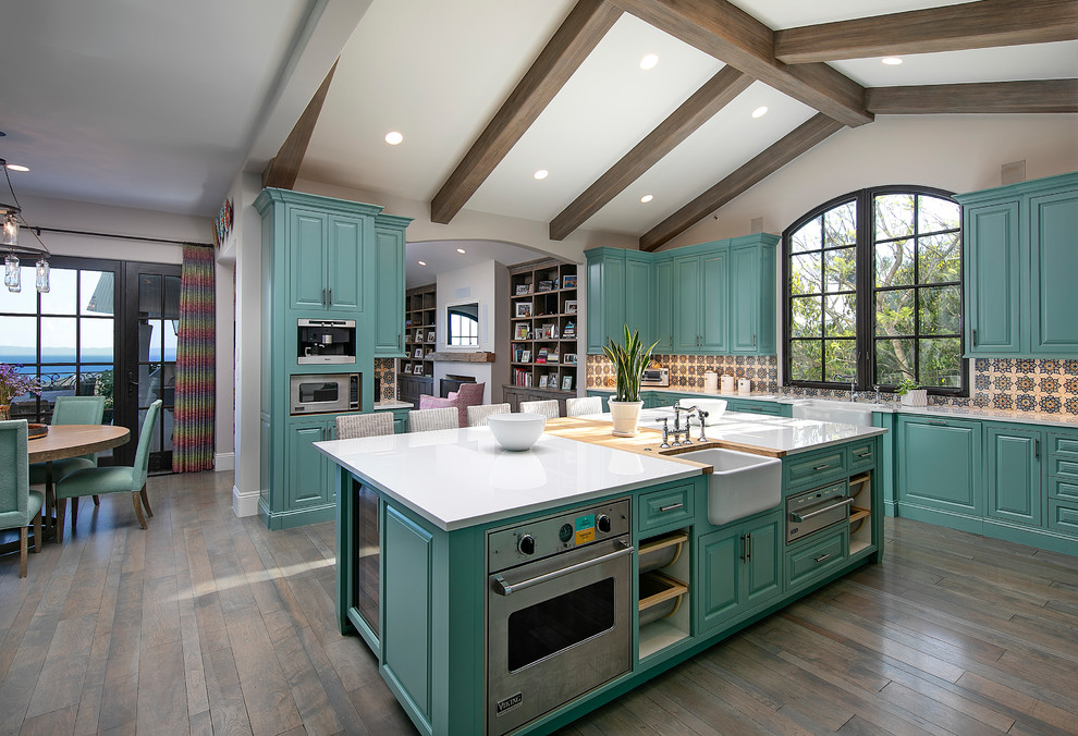 Design ideas for an expansive mediterranean l-shaped open plan kitchen in Santa Barbara with a farmhouse sink, raised-panel cabinets, turquoise cabinets, multi-coloured splashback, stainless steel appliances, medium hardwood floors, with island, brown floor and white benchtop.