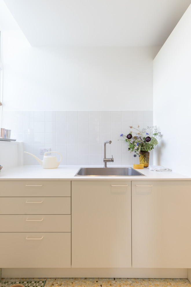 Design ideas for a large contemporary l-shaped dedicated laundry room in London with a drop-in sink, flat-panel cabinets, white cabinets, solid surface benchtops, white splashback, ceramic splashback, white walls, porcelain floors, a stacked washer and dryer, green floor and white benchtop.