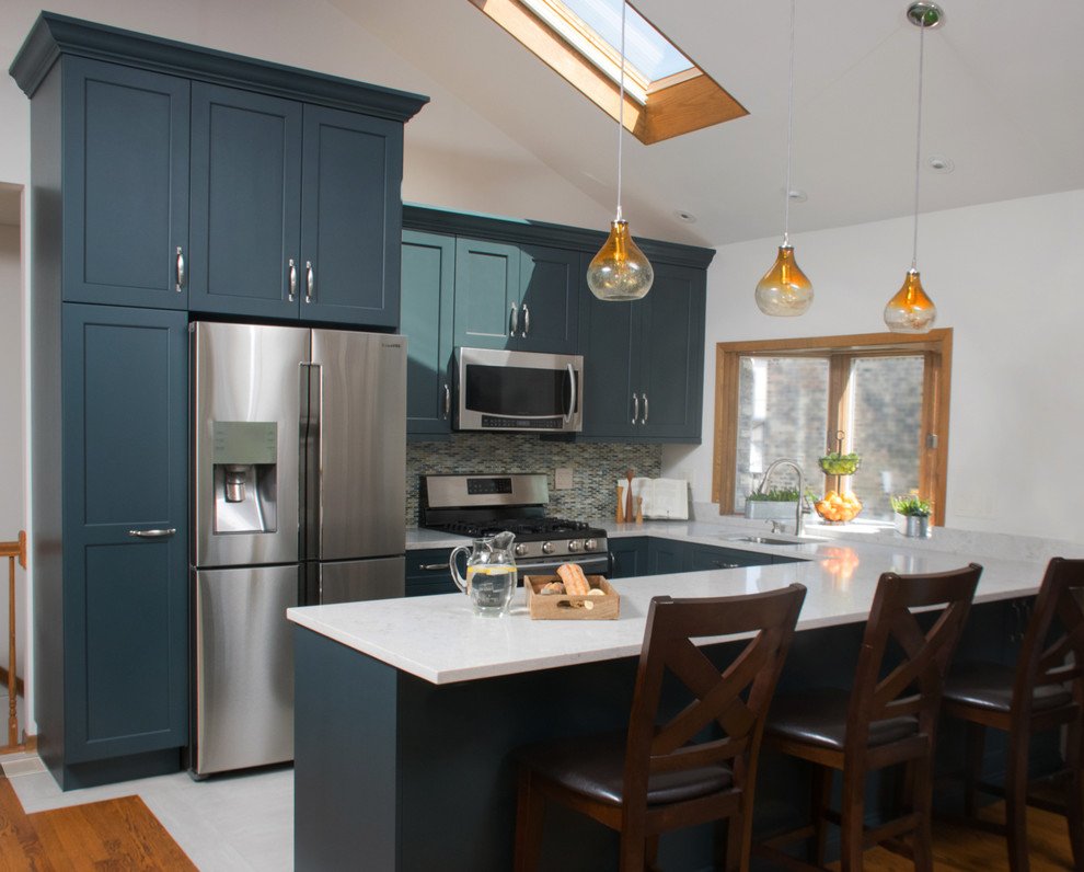 This is an example of a mid-sized transitional u-shaped open plan kitchen in Chicago with an undermount sink, shaker cabinets, blue cabinets, quartz benchtops, multi-coloured splashback, glass tile splashback, stainless steel appliances, porcelain floors, a peninsula and white floor.