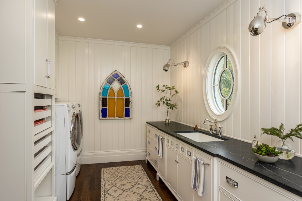 Inspiration for a mid-sized transitional galley dedicated laundry room in Other with an undermount sink, beaded inset cabinets, white cabinets, soapstone benchtops, white walls, dark hardwood floors, a side-by-side washer and dryer, brown floor and black benchtop.