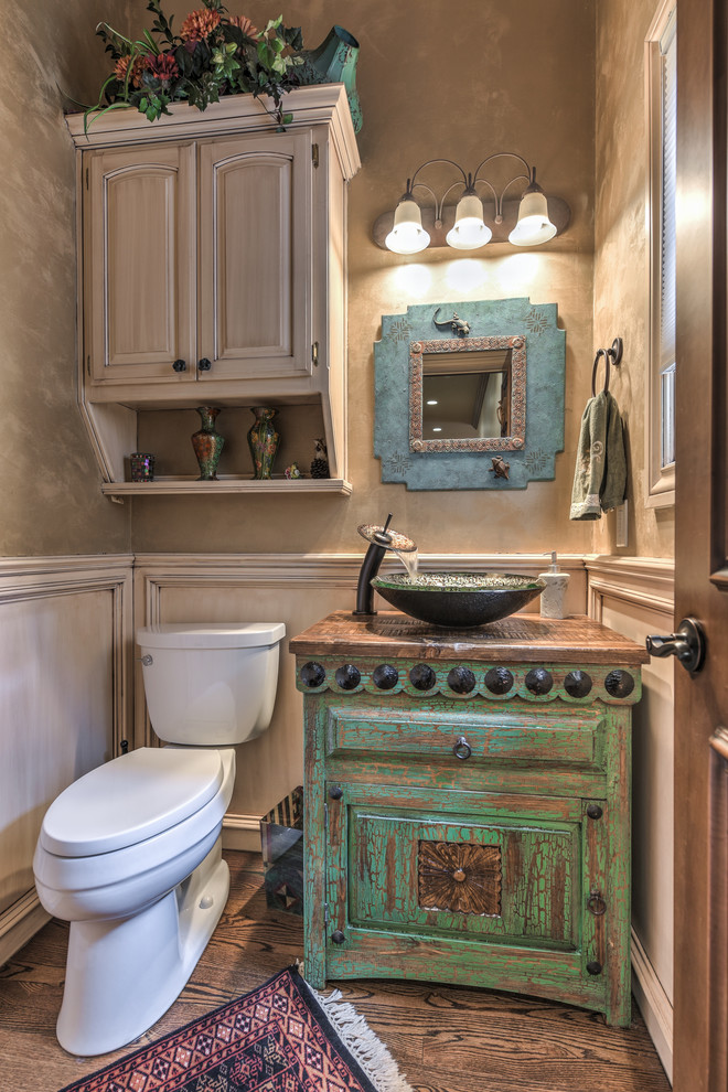 Photo of a mid-sized powder room in Oklahoma City with recessed-panel cabinets, distressed cabinets, a two-piece toilet, beige walls, dark hardwood floors, a vessel sink, wood benchtops and brown floor.