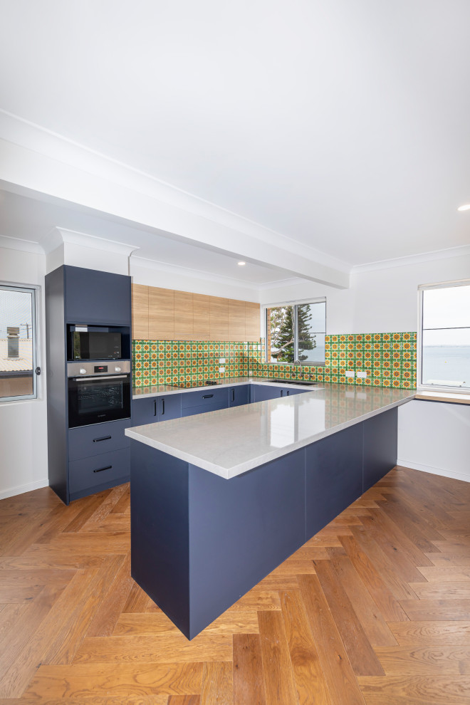 This is an example of a small midcentury u-shaped kitchen pantry in Central Coast with a drop-in sink, blue cabinets, multi-coloured splashback, black appliances, light hardwood floors, with island and grey benchtop.