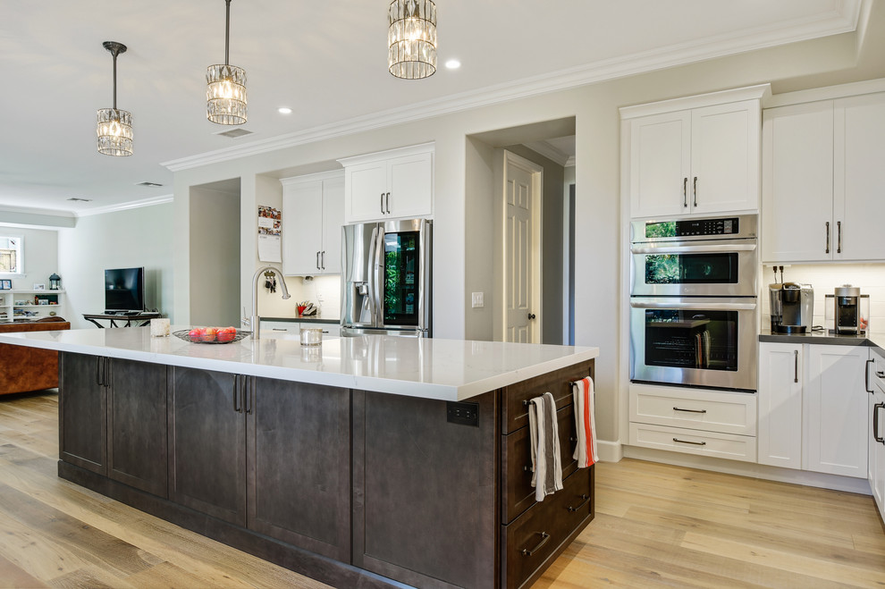 Photo of a large contemporary l-shaped open plan kitchen in San Diego with a farmhouse sink, shaker cabinets, white cabinets, quartz benchtops, white splashback, ceramic splashback, stainless steel appliances, light hardwood floors, with island, brown floor and white benchtop.