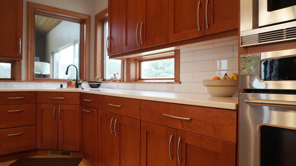 Large transitional l-shaped medium tone wood floor home design photo with an undermount sink, recessed-panel cabinets, brown cabinets, white backsplash, subway tile backsplash, stainless steel appliances, an island and white countertops