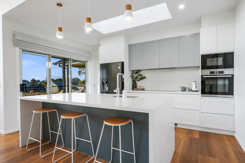 Photo of a mid-sized contemporary galley open plan kitchen in Other with an undermount sink, shaker cabinets, white cabinets, quartz benchtops, white splashback, black appliances, vinyl floors, with island, brown floor, white benchtop and stone slab splashback.