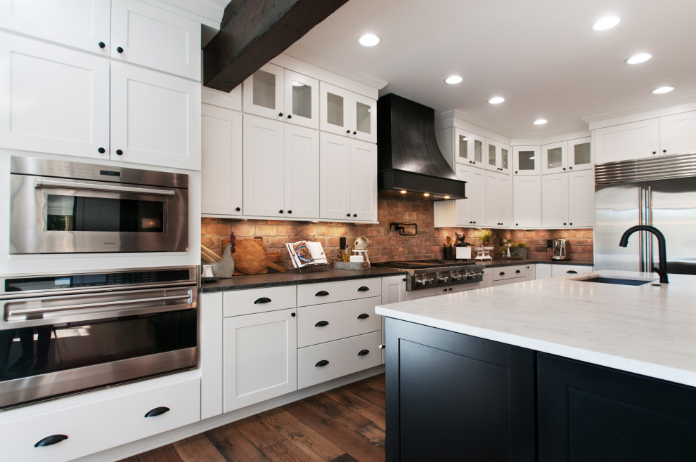 This is an example of a large country u-shaped open plan kitchen in Nashville with an undermount sink, shaker cabinets, white cabinets, quartz benchtops, multi-coloured splashback, brick splashback, stainless steel appliances, medium hardwood floors, with island, brown floor, white benchtop and exposed beam.