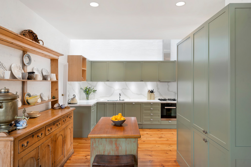This is an example of a mid-sized transitional l-shaped eat-in kitchen in Melbourne with a drop-in sink, shaker cabinets, green cabinets, quartz benchtops, white splashback, stone slab splashback, black appliances, medium hardwood floors, with island, brown floor and white benchtop.