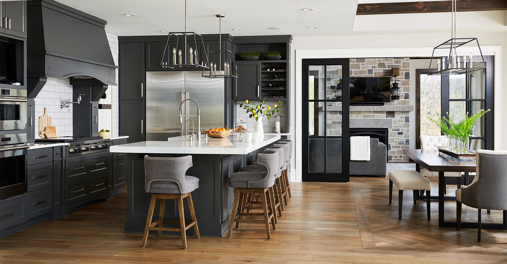 This is an example of a large transitional l-shaped eat-in kitchen in Minneapolis with medium hardwood floors, brown floor, an undermount sink, shaker cabinets, black cabinets, quartz benchtops, white splashback, subway tile splashback, stainless steel appliances, with island and white benchtop.