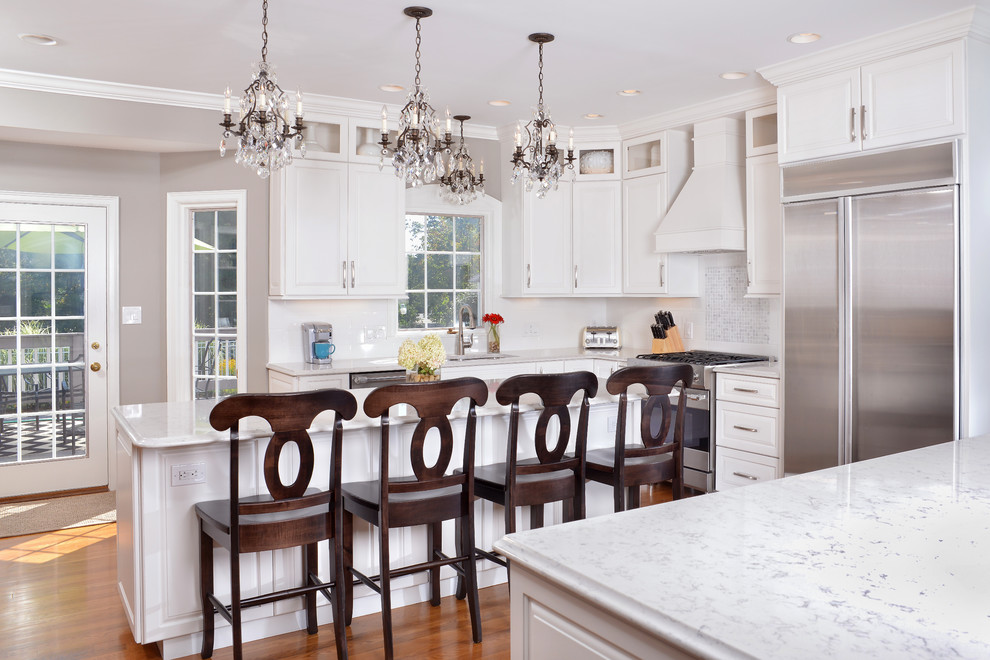 Mid-sized elegant l-shaped medium tone wood floor and brown floor kitchen photo in Cleveland with a double-bowl sink, raised-panel cabinets, white cabinets, quartz countertops, white backsplash, ceramic backsplash, stainless steel appliances and an island