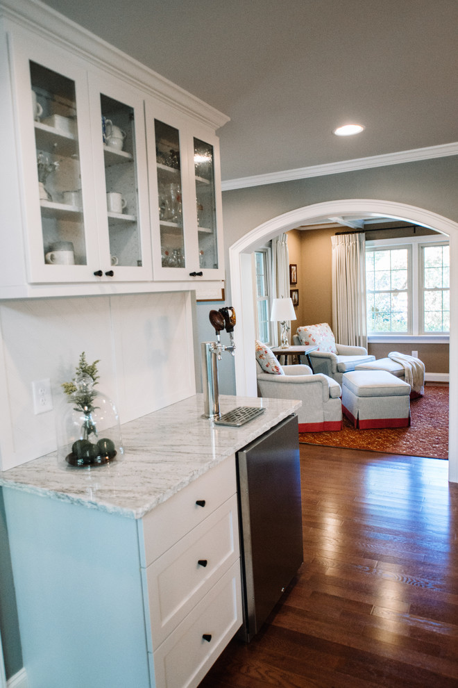 Large transitional l-shaped eat-in kitchen in Charlotte with a farmhouse sink, shaker cabinets, white cabinets, granite benchtops, white splashback, ceramic splashback, stainless steel appliances, medium hardwood floors, with island, brown floor and grey benchtop.