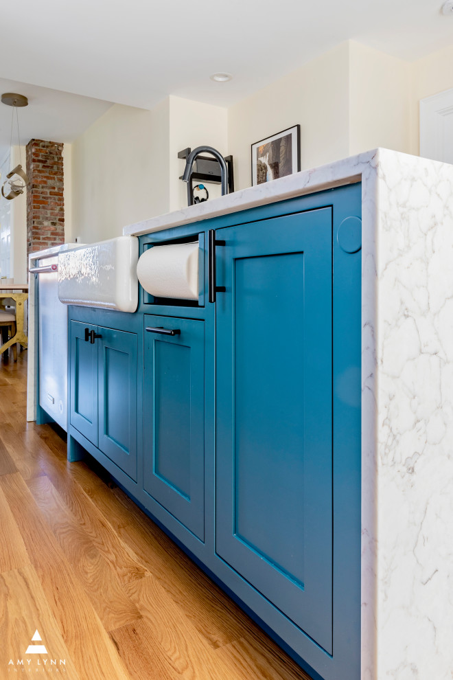 This is an example of a mid-sized eclectic single-wall eat-in kitchen in Boston with a farmhouse sink, shaker cabinets, blue cabinets, quartz benchtops, white splashback, ceramic splashback, stainless steel appliances, light hardwood floors, with island, brown floor and white benchtop.