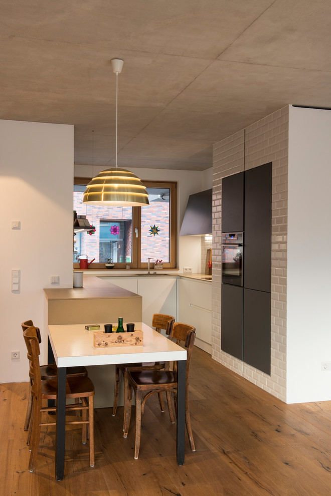 Photo of a small contemporary u-shaped open plan kitchen in Cologne with a drop-in sink, flat-panel cabinets, white cabinets, white splashback, subway tile splashback, black appliances, medium hardwood floors, no island and brown floor.