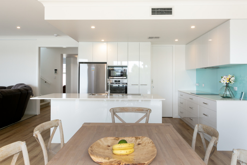 Photo of a large beach style l-shaped open plan kitchen in Adelaide with a drop-in sink, white cabinets, quartz benchtops, blue splashback, glass sheet splashback, stainless steel appliances, light hardwood floors, with island, multi-coloured floor and white benchtop.