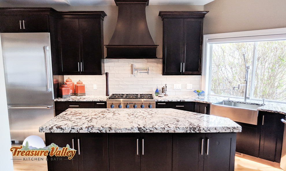 Example of a mid-sized transitional l-shaped light wood floor open concept kitchen design in Boise with a farmhouse sink, shaker cabinets, granite countertops, glass tile backsplash, stainless steel appliances and an island