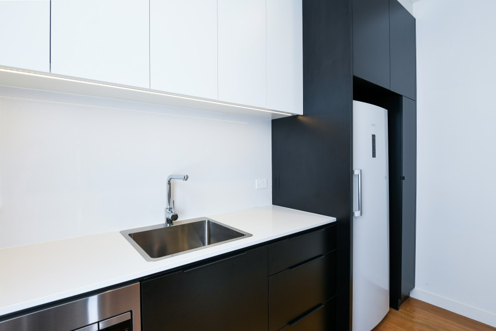 This is an example of a large modern galley open plan kitchen in Sydney with a drop-in sink, flat-panel cabinets, black cabinets, quartz benchtops, white splashback, stone slab splashback, black appliances, medium hardwood floors, brown floor and white benchtop.