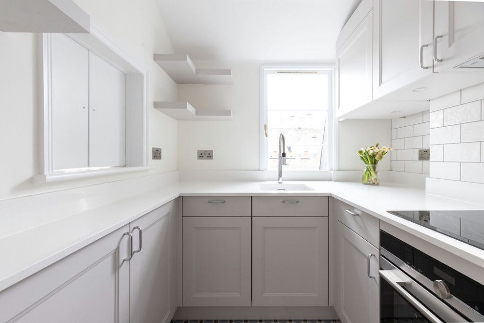 Small modern grey and white open plan kitchen in London with grey cabinets, white splashback, ceramic splashback, stainless steel appliances, no island, multi-coloured floors and white worktops.