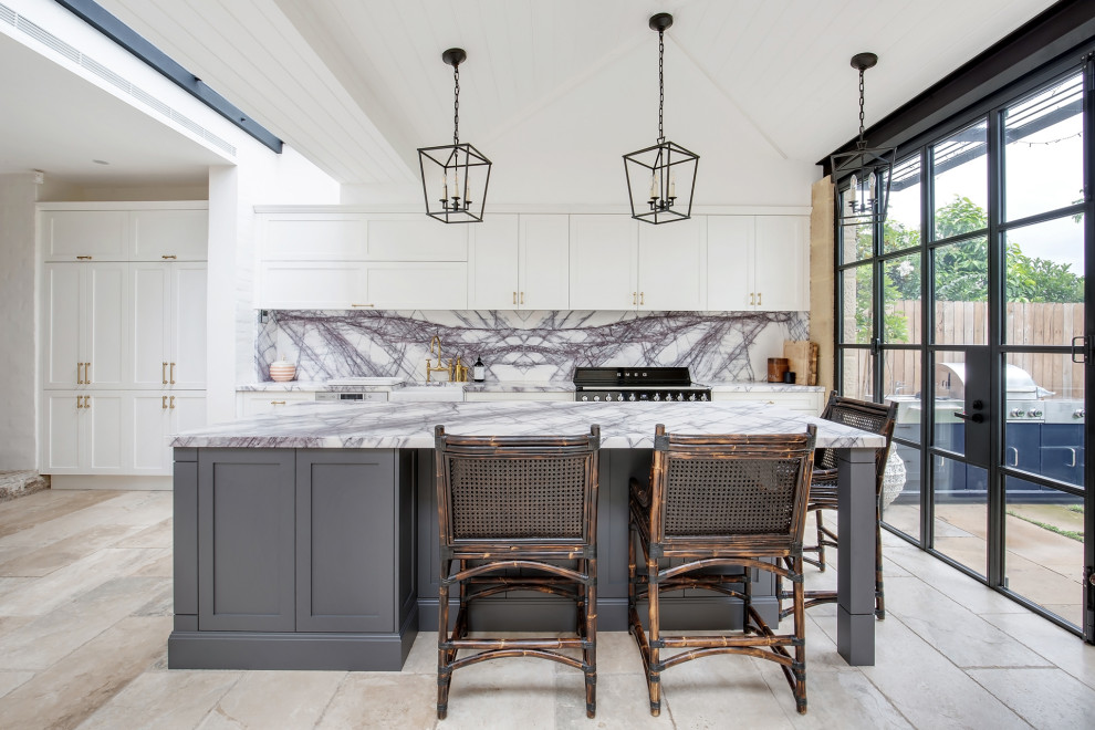 This is an example of an expansive transitional galley open plan kitchen in Sydney with a farmhouse sink, shaker cabinets, white cabinets, grey splashback, black appliances, with island, beige floor, grey benchtop, timber, vaulted, marble benchtops, marble splashback and travertine floors.