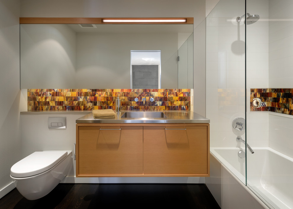 Photo of a mid-sized contemporary master bathroom in Portland with medium wood cabinets, an alcove tub, a shower/bathtub combo, a wall-mount toilet, orange tile, glass tile, white walls, dark hardwood floors, an integrated sink, stainless steel benchtops, black floor, a hinged shower door and flat-panel cabinets.