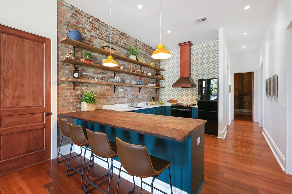 Industrial u-shaped eat-in kitchen in New Orleans with an undermount sink, recessed-panel cabinets, blue cabinets, wood benchtops, multi-coloured splashback, brick splashback, black appliances, medium hardwood floors, a peninsula, brown floor and white benchtop.