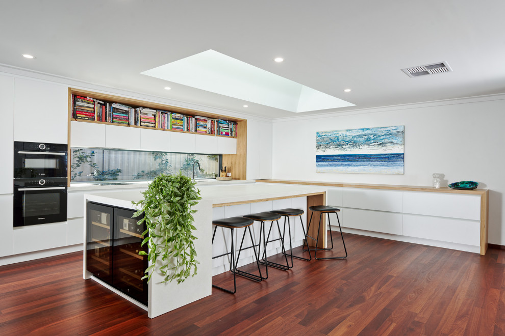 Inspiration for a large modern l-shaped eat-in kitchen in Perth with an undermount sink, flat-panel cabinets, white cabinets, quartz benchtops, white splashback, window splashback, black appliances, dark hardwood floors, with island, brown floor and white benchtop.