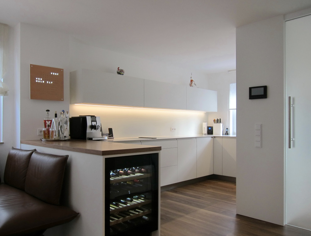 This is an example of a medium sized contemporary u-shaped open plan kitchen in Cologne with a built-in sink, flat-panel cabinets, white cabinets, engineered stone countertops, white splashback, glass sheet splashback, black appliances, vinyl flooring, a breakfast bar, brown floors and beige worktops.