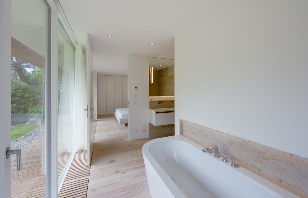This is an example of a large contemporary master bathroom in Berlin with flat-panel cabinets, white cabinets, a drop-in tub, beige tile, stone slab, white walls, light hardwood floors and an integrated sink.