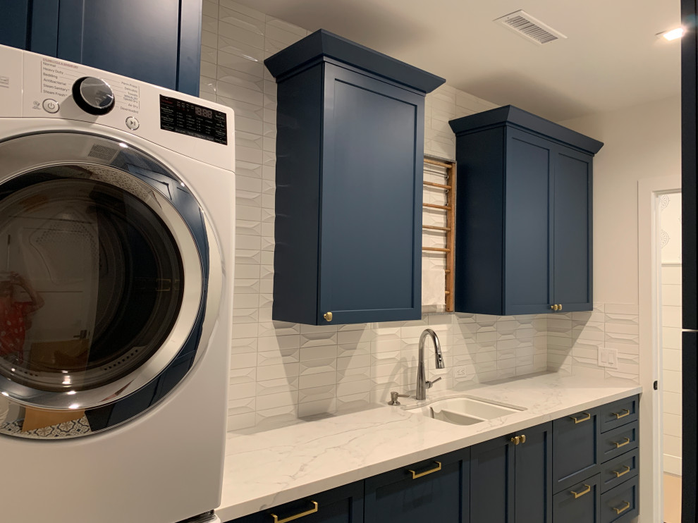 Inspiration for a mid-sized coastal galley ceramic tile and multicolored floor dedicated laundry room remodel in Orange County with a double-bowl sink, shaker cabinets, blue cabinets, quartz countertops, white backsplash, ceramic backsplash, white walls, a stacked washer/dryer and white countertops