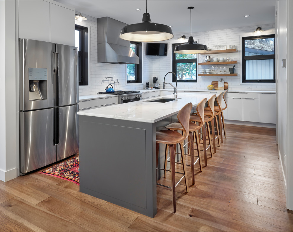 This is an example of a midcentury l-shaped kitchen in Edmonton with an undermount sink, flat-panel cabinets, white cabinets, white splashback, subway tile splashback, stainless steel appliances, medium hardwood floors, with island and white benchtop.